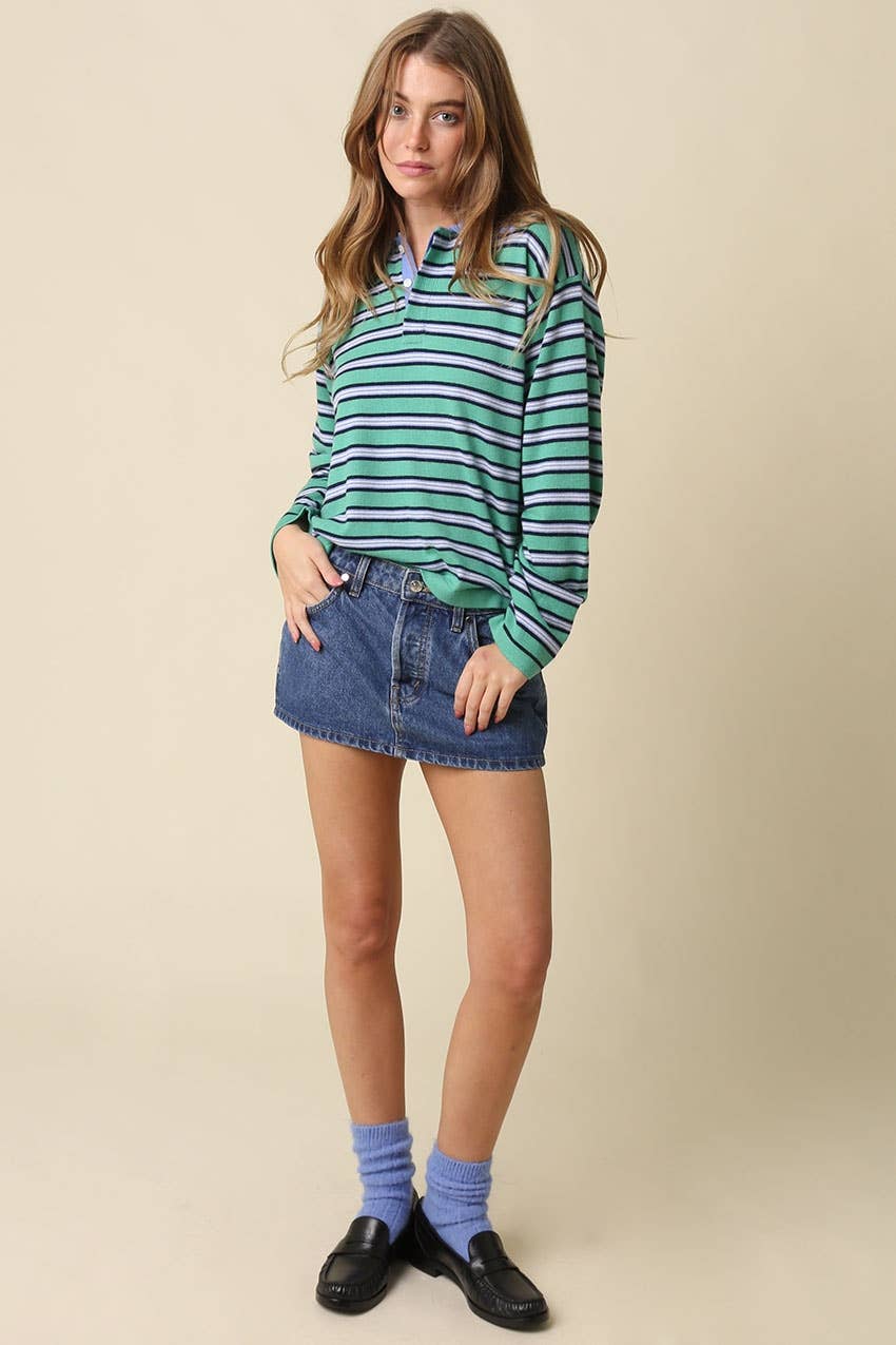 A woman modeling a green and white striped long-sleeve top with a high collar, paired with a denim mini skirt, blue socks, and black loafers, against a solid beige studio background.