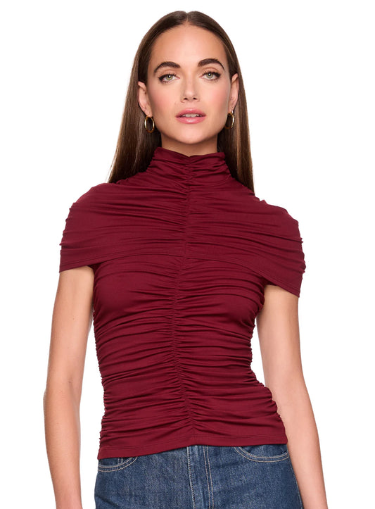 A woman modeling a short-sleeve ruched mock neck top in deep burgundy, paired with blue jeans, against a plain white background.