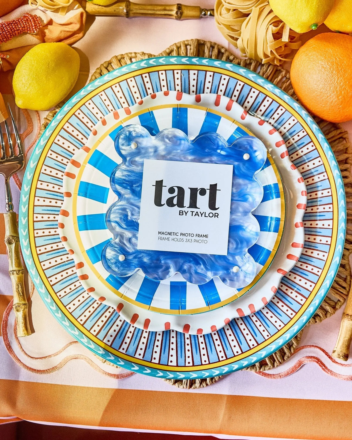 A decorative round magnetic photo frame with a blue and white striped center and a turquoise rim, styled on a table with lemons, pasta, and gold-toned flatware. The center card reads 'tart BY TAYLOR'.