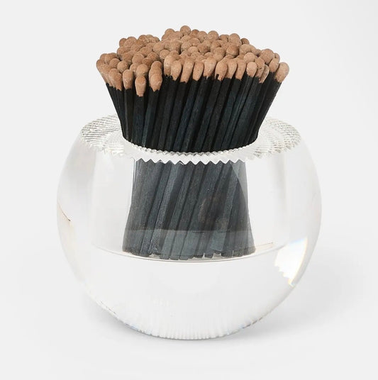 A clear glass bowl filled with black matches topped with tan wooden tips, photographed on a clean white background.