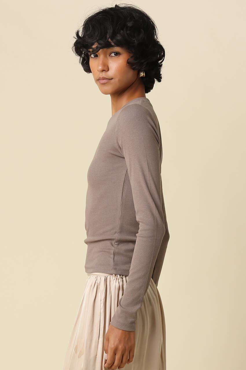 A woman with short dark curly hair is shown in profile wearing a fitted long-sleeve top in a muted taupe-brown color and a light beige skirt, against a plain warm beige studio background.