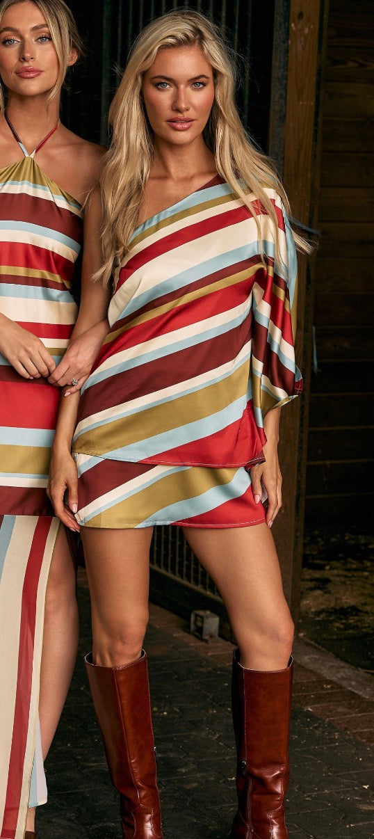 A woman models a one-shoulder mini dress with diagonal stripes in red, mustard yellow, light blue, and cream, paired with brown knee-high boots. Another model is partially visible on the left.