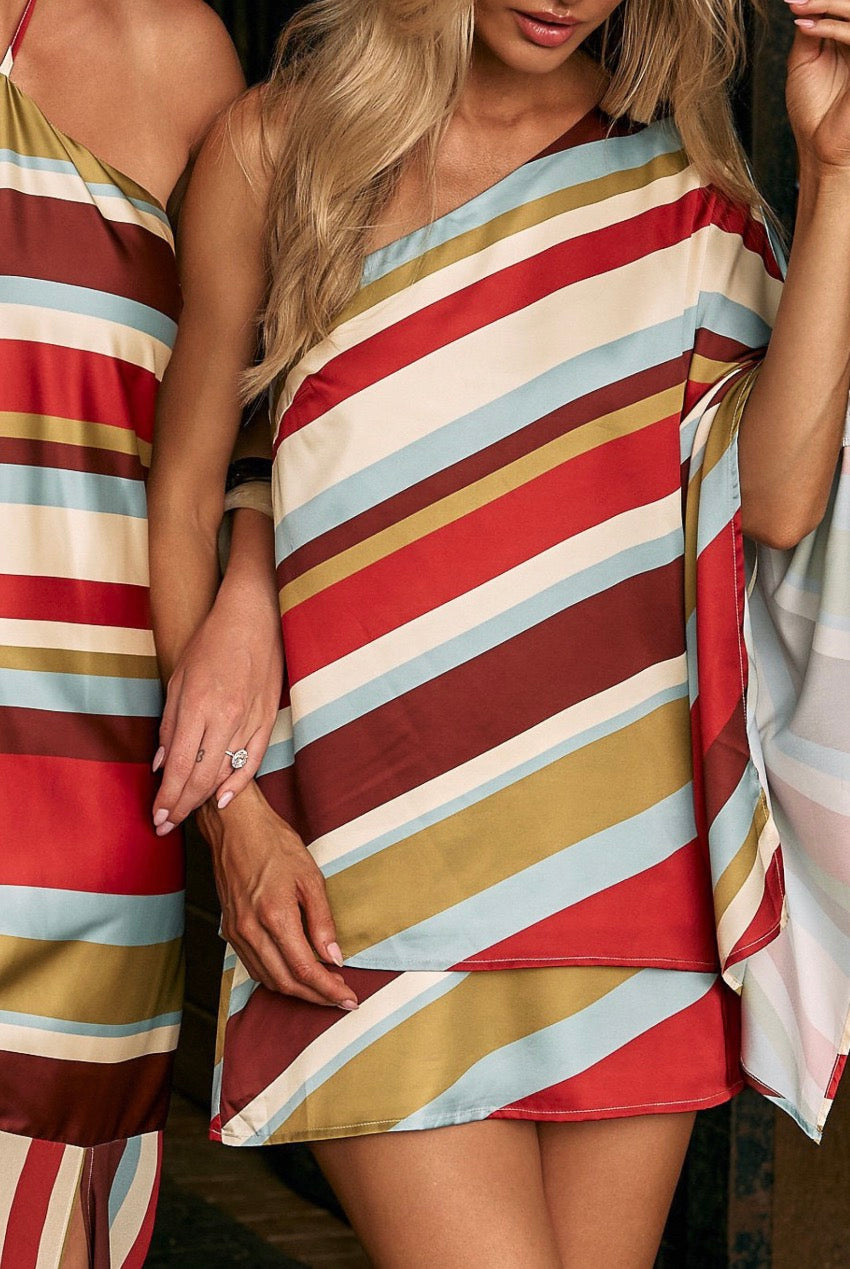 A woman wearing a short, one-shoulder satin slip dress with diagonal stripes in red, cream, light blue, mustard yellow, and burgundy.