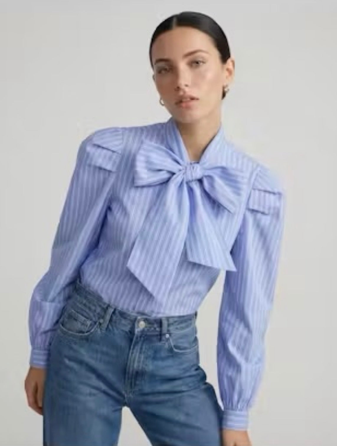A woman wearing a light blue and white vertically striped blouse with a large bow at the collar and blue jeans, photographed against a plain light gray studio background.