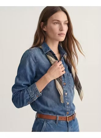 A woman wearing a blue denim button-up shirt with rolled sleeves, a brown belt, and a patterned scarf, photographed against a plain light background.