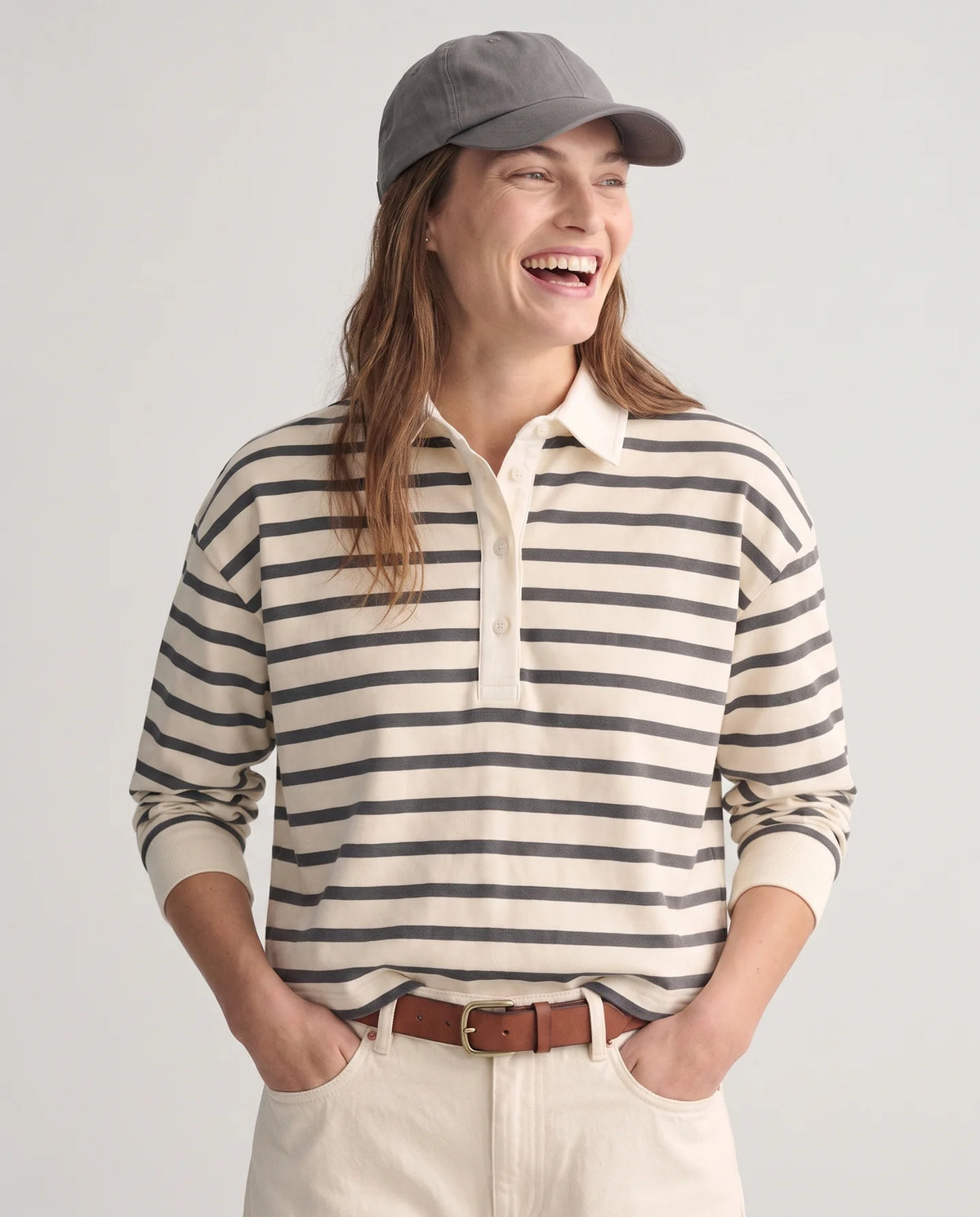 A woman wearing a cream and dark gray horizontally striped long-sleeve polo shirt, cream pants, a brown belt, and a gray baseball cap, smiling against a light gray studio background.