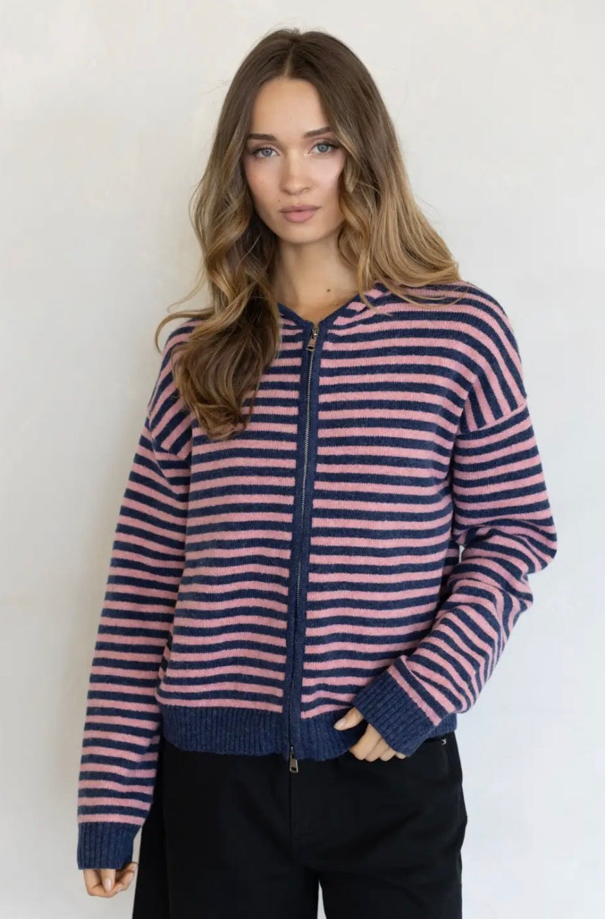 A woman wearing a navy and pink striped zip-up sweater with dark navy ribbed trim, paired with black pants, standing against a plain white background.