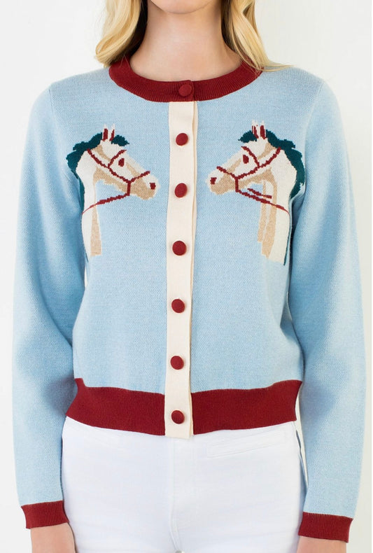 A light blue knit cardigan sweater with a cream center placket and red trim, featuring two embroidered carousel horse motifs on the chest. The cardigan is shown on a model wearing white pants against a plain white background.