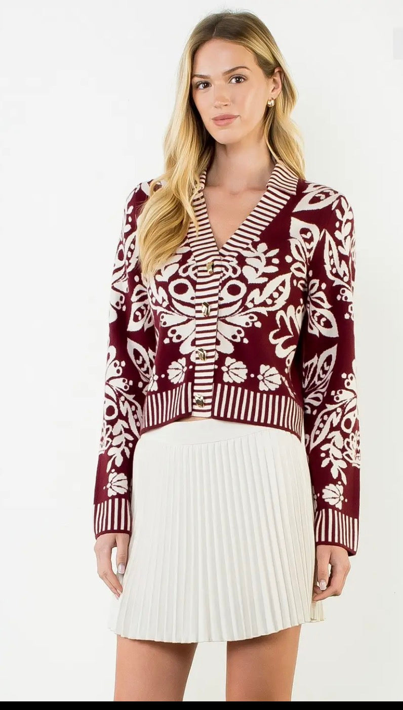 A woman modeling a burgundy and white patterned knit cardigan with a V-neck, paired with a white pleated mini skirt, against a plain white background.
