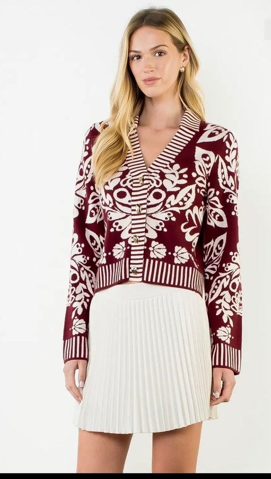 A woman modeling a burgundy and white patterned knit cardigan with a V-neck, paired with a white pleated mini skirt, against a plain white background.