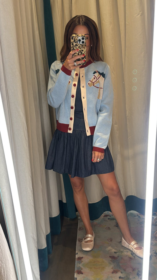 A woman takes a mirror selfie in a fitting room wearing a light blue varsity-style jacket with red and gold trim, a dark navy pleated mini skirt, and white loafers.