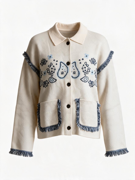 An off-white cropped cardigan jacket featuring blue embroidered paisley and floral patterns, fringed trim along the collar, cuffs, hem, and pockets, and dark buttons down the front.