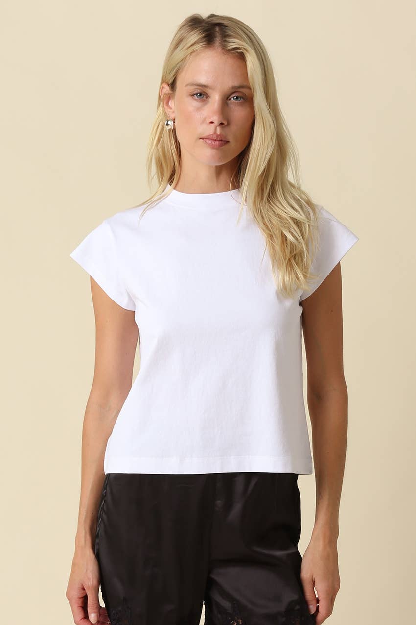 A blonde woman modeling a white short-sleeve crewneck t-shirt with black pants, standing against a solid beige background.