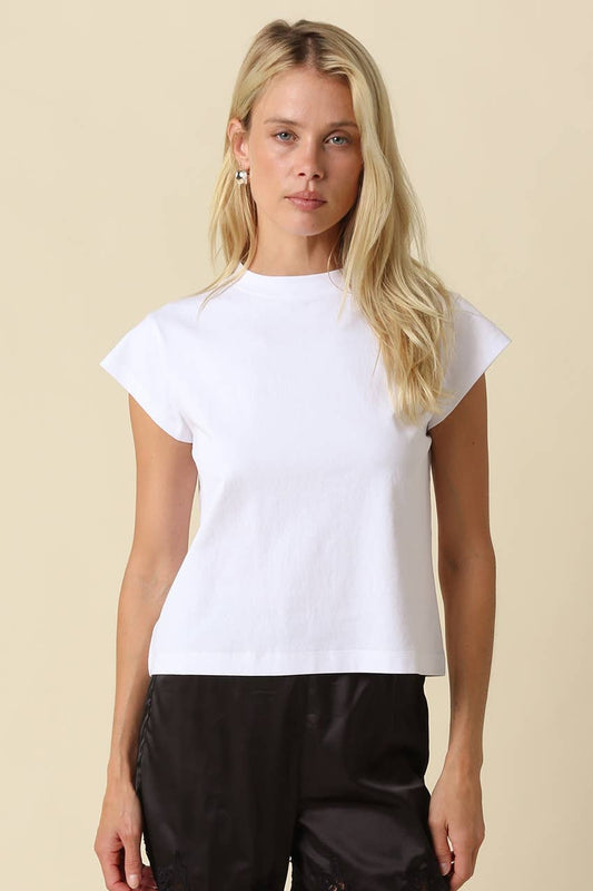 A blonde woman modeling a white short-sleeve crewneck t-shirt with black pants, standing against a solid beige background.