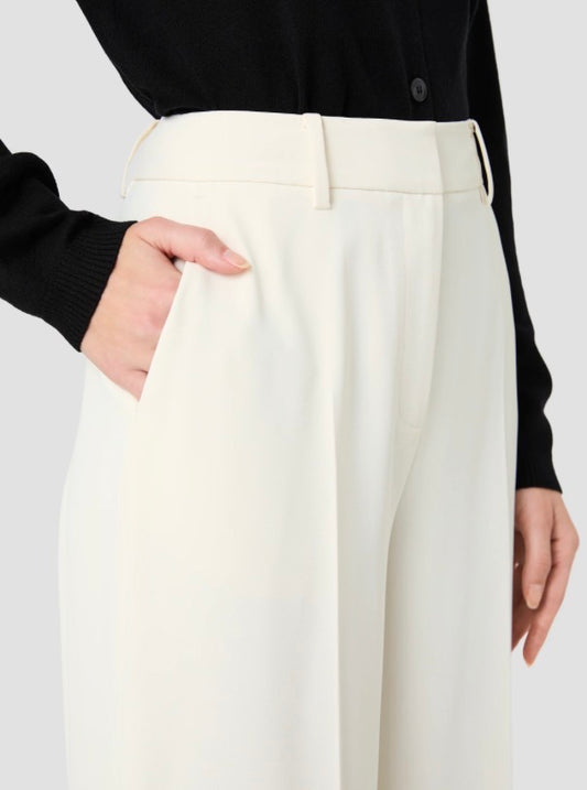 A close-up of a model wearing high-waisted, wide-leg trousers in off-white with a matching belt, paired with a black long-sleeve top against a light gray background.