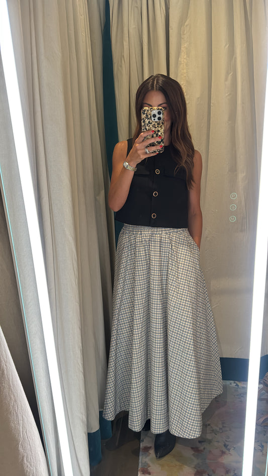A woman takes a mirror selfie in a fitting room wearing a sleeveless black button-front top and a high-waisted, full-length A-line skirt with a small-scale black and white gingham pattern.